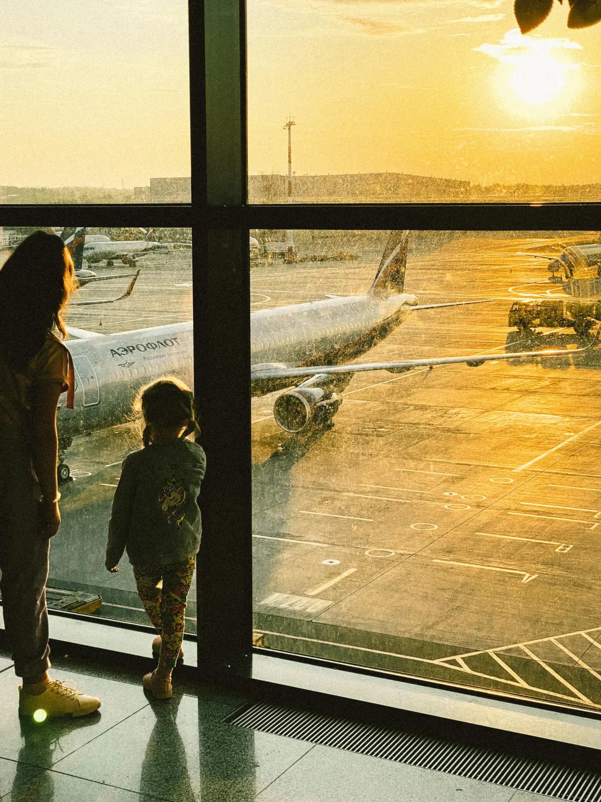Silhuetter av en voksen og et barn som ser opp mot fly ved soloppgang på en flyplass, gyllent lys